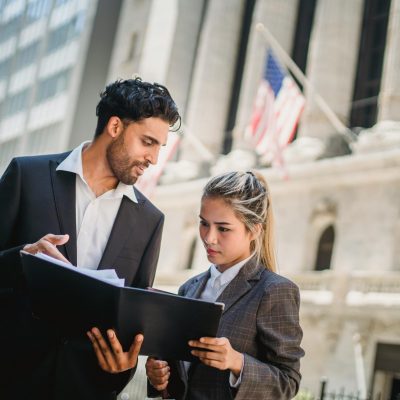 Two professionals in suits discussing documents outside a corporate building in daylight.