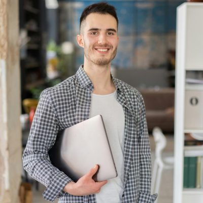 Cheerful young adult man holding a laptop, standing in a contemporary office space.
