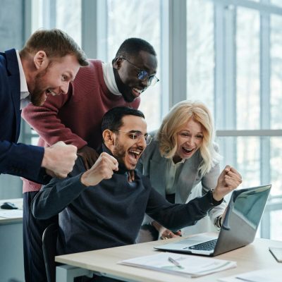 A diverse team in an office celebrates a successful achievement while looking at a laptop.