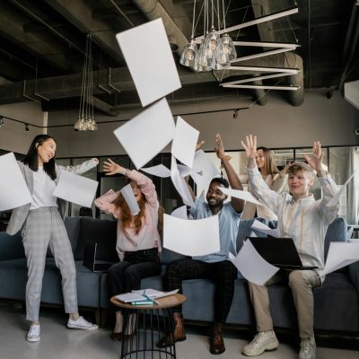 A diverse team of adults celebrates success with papers flying in an office setting.